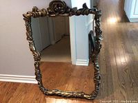 Front view of the gold-toned decorative wall mirror with carved floral details on the frame, standing on a hardwood floor.