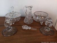 Crystal and glassware assortment including two covered bowls, a pitcher, two candle holders, and a glass stirrer arranged on a wooden surface.
