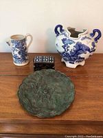 Photo showing the entire lot of four decorative items placed on a wooden surface, including two blue and white ceramic pieces, a bronze dish, and a small decorated box.