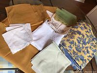 Overview of various linens and fabrics arranged on a table including gold drapery panels, white napkins, a floral piece, and other linens.