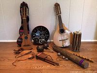 Overview of all instruments arranged on wooden floor: charango, handpan, cylinder drum, rainstick, panpipes, flute, maracas, bells, claves.