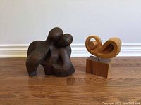 Photo showing two wooden abstract sculptures on floor: dark brown embracing figures and light smooth flowing shape on base