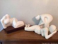Three white plaster sculptures displayed on a wooden surface against white wall, showing different abstract human poses.