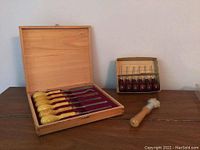 Opened wooden case showing 8 wood carving chisels with yellow wooden handles inside, red felt lining, small separate box of 6 chisels with dark wooden handles and wooden cleaning brush in front.