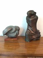 Image showing two sculptures on wooden surface: a taller dark brown twisted form and a smaller lighter rounded form on wooden base