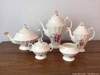 Full tea set displayed on wooden table including tall coffee pot, teapot, sugar bowl with lid, creamer, and small covered sugar pot. They feature white embossed porcelain with pink rose floral detail.