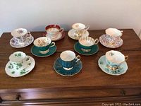 View of full set of 8 teacups and saucers arranged on wooden surface showing different floral patterns and gold trim.