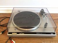 Front view showing entire Technics turntable with clear dust cover on a wood floor with power cord visible.