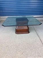 View showing square clear glass top with beveled edges on wooden pedestal base, placed on concrete floor in front of a blue rolling door.