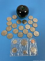 Photo showing spread of twenty-one 1968 silver dollar coins around a green glass apple for size reference, with the sealed mint coin set displayed below.