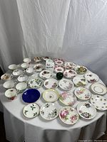 Wide-angle photo showing all cups and saucers arranged on a round white table with a small green apple for size reference. Various floral patterns and some gold trim visible. All items laid out for clear view.