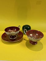 Three porcelain items displayed with green apple for scale: one cup with saucer on left, one orphan cup on right
