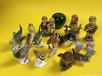 Side view of 15 porcelain figurines including birds, children Hummel figurines, and three Herend miniature cat figurines for size reference with green apple.