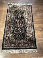 Full view of rectangular black and red silk rug with cream fringe on hardwood floor.