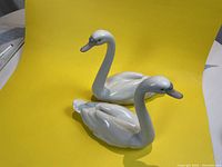 Front angled view of two white porcelain swan figurines on a yellow background showing size and glossy finish