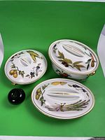 Top view of three Royal Worcester stoneware covered oven dishes with naturalistic fruit and vegetable designs alongside a green apple for size reference.