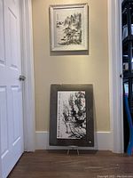 Photo showing two paintings, one framed hanging on the wall and one unframed resting on the floor against the wall.