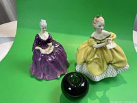 Two Royal Doulton figurines on green background next to green apple for size reference. One in yellow dress holding letter, one in purple dress holding dog.