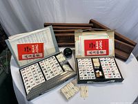 Overall lot view: two open Mahjong cases with tiles and counters on a table, wooden tile racks behind