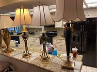 Three metal table lamps on kitchen counter, two with twisted gold bases and square beige shades, third with black base and scalloped beige shade.