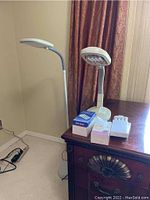 Photo shows two lamps: a floor lamp with a slim curved silver neck and circular LED light head, and a white desk lamp with rectangular fluorescent light head. Several packs of fluorescent replacement bulbs are placed next to the desk lamp on a wooden surface.