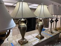 Two identical metal table lamps with fabric shades on a granite countertop in a kitchen. Lamps have urn-style bases with dual handles and square pedestal feet. Shades are bell-shaped and light-colored.