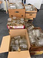 Twelve cardboard boxes filled with various sizes of clear glass canning jars, some jars stacked inside others, no lids visible, dusty and requiring cleaning.