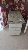 White nightstand with three drawers, round knobs, and granite top.