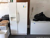 Front view of white Kenmore side-by-side refrigerator and freezer next to a chest freezer, showing the external ice and water dispenser on the left door.