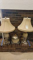 Pair of decorative lamps with beige shades and matching decorative box set on a mantel against a brick wall.