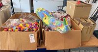 Two cardboard boxes filled with assorted luau party supplies including multicolored leis and an inflatable tropical item.