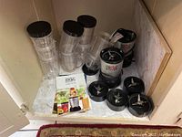 Two Magic Bullet blender bases with multiple transparent blending cups stacked behind them, blade attachments arranged in front, and user guides placed nearby in a cabinet.