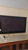 Front view of Panasonic 64 inch plasma TV on granite countertop showing the full screen, frame, and remote control with cables below.