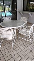 Full set of patio furniture showing round frosted glass tabletop and four white plastic webbed chairs on tiled floor.