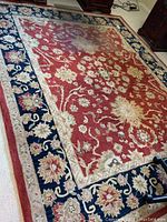 Full view of rectangular floral area rug with red center and blue border on light carpeted floor.