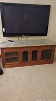 Wide front view of wooden entertainment cabinet with mounted TV above, showing removable granite or marble top, glass paneled front doors, and wood detailing.