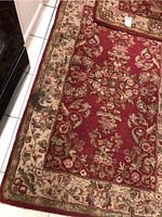 Close-up photo of one wool rug showing the detailed floral pattern on red field with beige border, laid on white tiled floor.