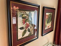 Photo showing two framed botanical prints hanging on a wall, depicting flowers and leaves with dark frames and burgundy mats.