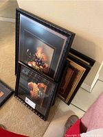 Photo showing a stack of five framed wall art pieces leaning against a wall, featuring fruit still life paintings including grapes and peaches.
