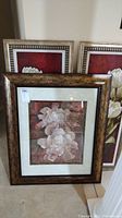 Framed floral wall art arranged with two matching red-background pictures standing behind a larger framed picture with white flowers and matted frame.
