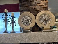 Photo showing set of two round decorative plates and two tall metal candle stands with ornate scrollwork and beige pillar candles on mantel with brick backdrop.