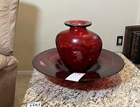 Red glass vase and decorative bowl set positioned on a granite surface next to furniture and wall outlet, highlighting color and dimensions.