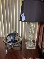 Photo of black pewter globe with geographic details supported by silver-toned circular frame and tripod legs, sitting on wooden surface next to clear acrylic lamp with black shade.