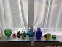 Full view of nine decorative glass items including vases, figurines, paperweight, and candle holder on window sill