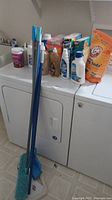 Photo showing several bottles and boxes of cleaning supplies on top of washer/dryer units, including Arm & Hammer baking soda, Oxi softener, Cheer detergent, dish soap, bleach plus, and a drain cleaner.