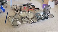 Wide view showing assortment of pots, pans, lids, and steamer basket on carpeted floor