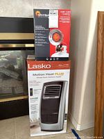 Photo showing boxed Mr. Heater Propane Tank Top Heater on top of boxed Lasko Motion Heat Plus ceramic heater next to a fireplace.