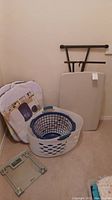 Two white plastic laundry baskets, six mesh laundry baskets packed, digital glass scale, and folded Lifetime table leaning against wall
