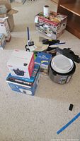 Lot of kitchen appliances shown on carpet including boxed and unboxed items, seen are Magefesa pressure cooker and Black & Decker rice cooker boxes.
