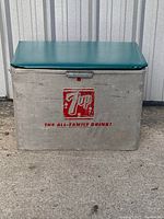 Front view of vintage 7UP metal cooler with green cushioned lid showing 7UP logo and slogan.
